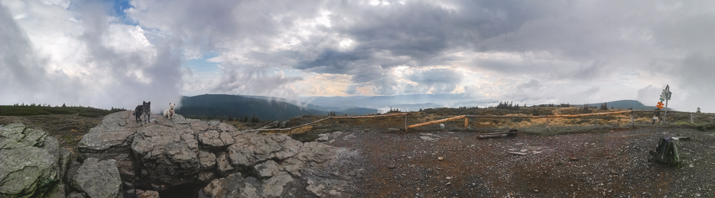 Panorama z góry Keprnik w Czechach. Na pierwszym planie formacje skalne, Fibi i Krakers. W tle górski pejzaż rozciągający się aż po horyzont. Mieszane chmury, jedne zwiastują deszcz, przez inne przebija się słońce i błękit nieba.